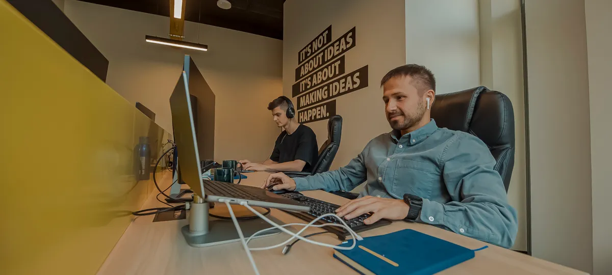 Twee developers werken aan een gedeeld bureau met monitoren, naast een muur met de slogan 'It's not about ideas. It's about making ideas happen.'